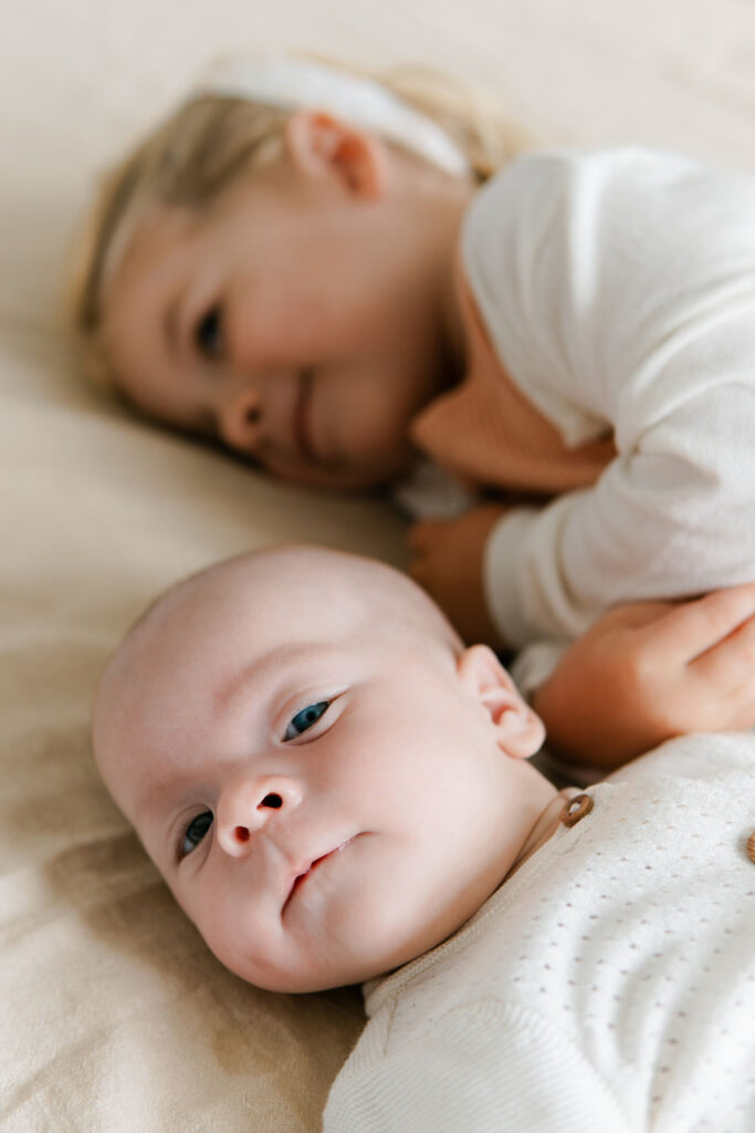 Baby kijkt in de camera en op de achtergrond ligt haar zus.