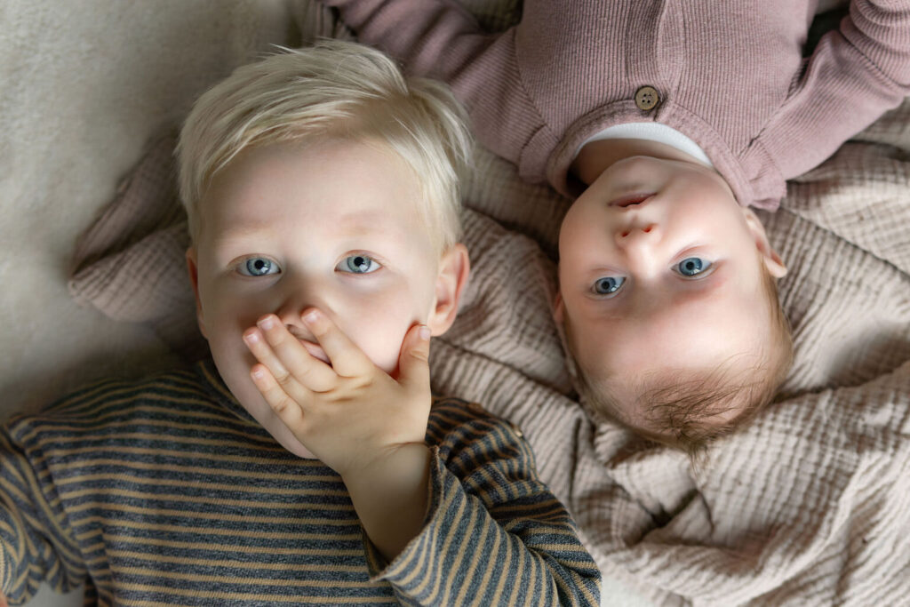 Baby en broer liggen op de grond op een kleed. Broer heeft hand voor de mond. 