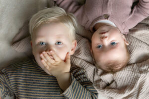 Baby en broer liggen op de grond op een kleed. Broer heeft hand voor de mond.