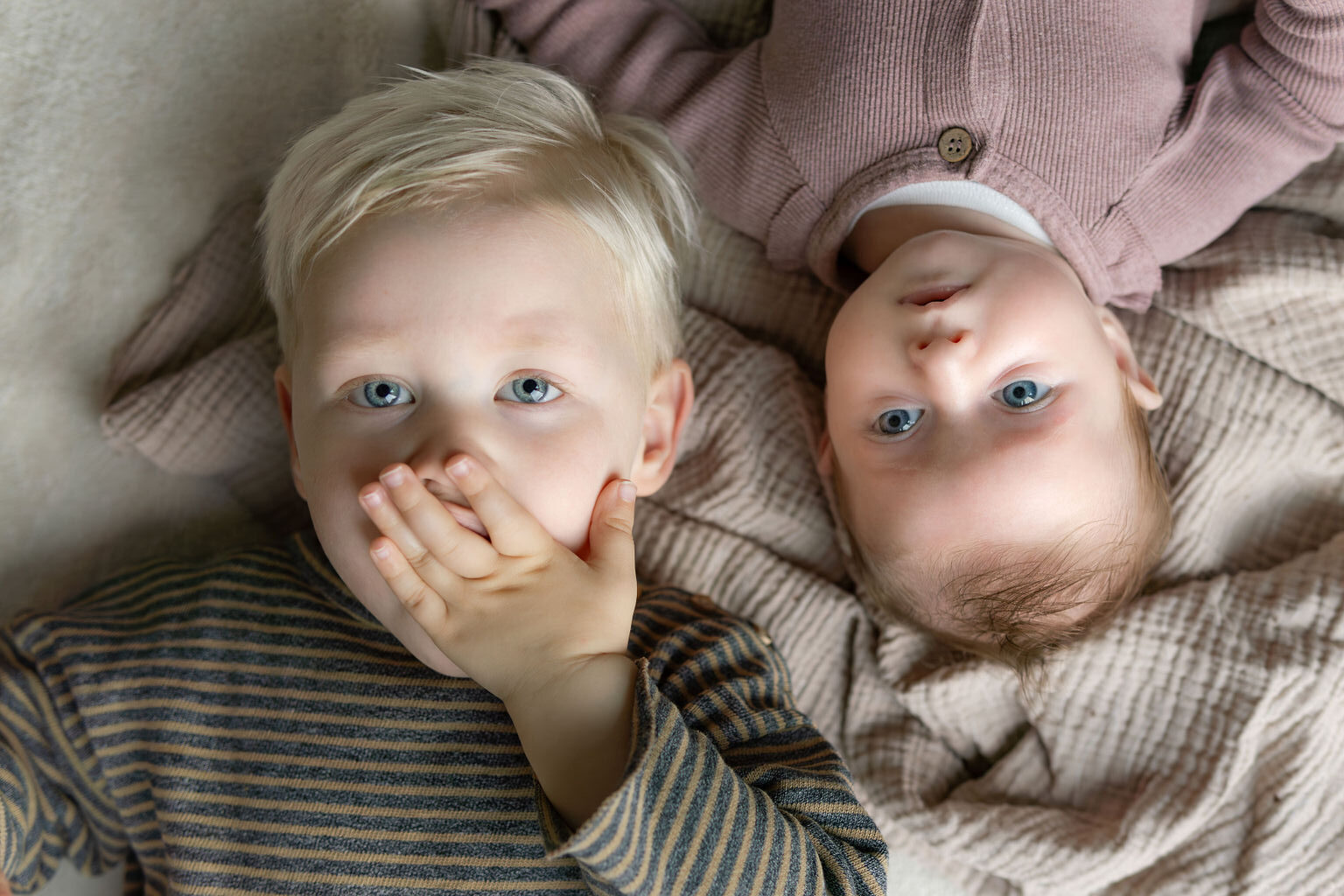 Baby en broer liggen op de grond op een kleed. Broer heeft hand voor de mond.
